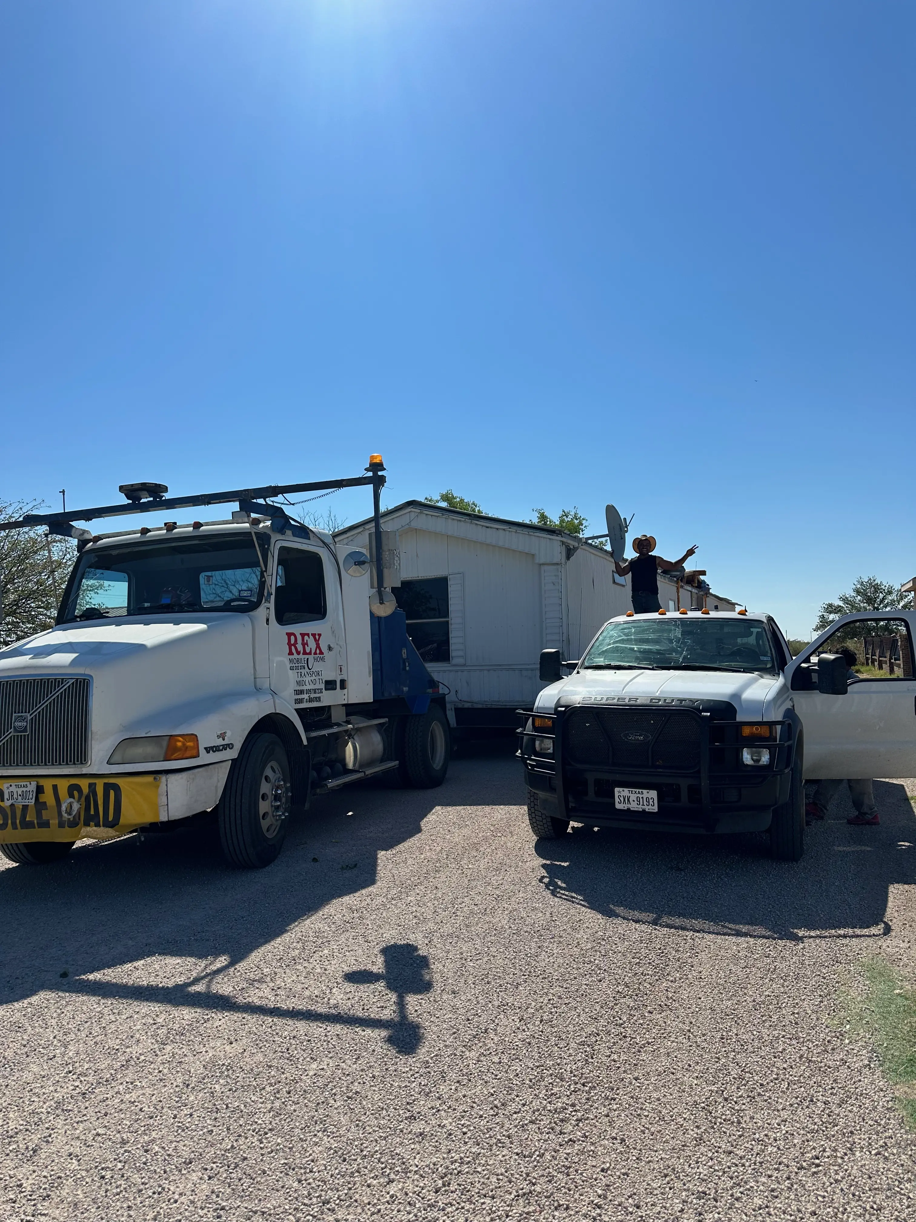 Commercial truck hauling a mobile home and a white truck with a man standing in the bed of the truck waving to the camera.